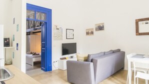 Ein modernes Wohnzimmer mit grauem Sofa, Fernseher, Esstisch mit Stühlen und einer blauen Tür, die zum Schlafzimmer führt.