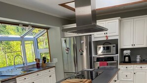 Une cuisine moderne avec des meubles blancs, des appareils en acier inoxydable, un grand îlot central doté d’une planche à découper et une vue sur la verdure environnante.