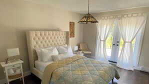 Une chambre à coucher avec une tête de lit capitonnée, un lit recouvert d’une courtepointe, une table de chevet avec une lampe, une chaise et une fenêtre avec des rideaux.