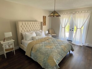 A bedroom with a tufted headboard, a bed with a quilt, a bedside table with a lamp, a chair, and a window with curtains.
