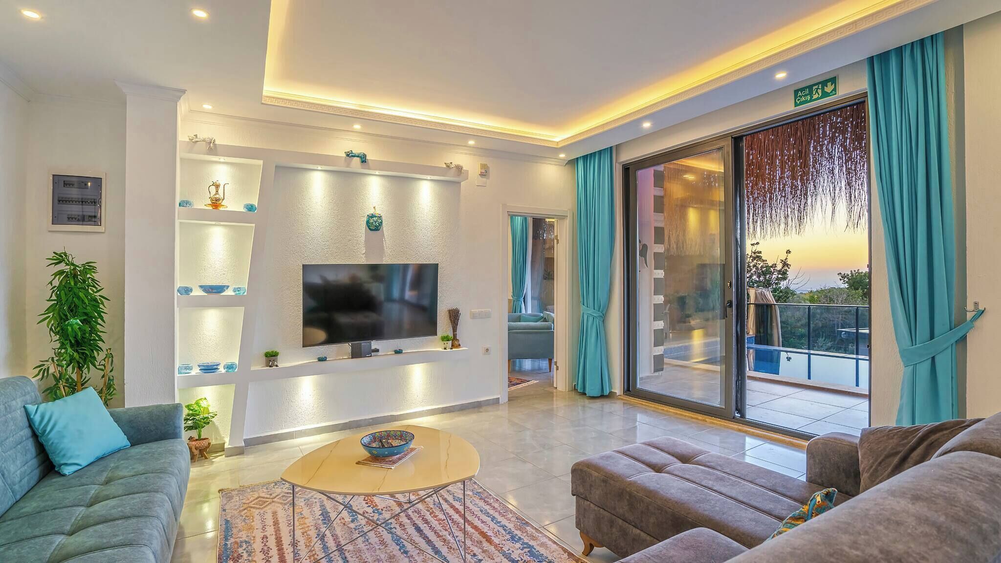 A modern living room with a flat-screen TV, a sofa, a coffee table, and a sliding glass door leading to an outdoor balcony.