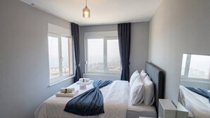 A modern hotel room with a large bed, two windows with curtains, a bedside table, and a view of the city. - Quiet and Comfortable in Sinop Karakum Site 2 (Sinop Merkez)