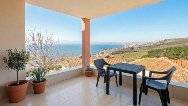 Ein Balkon mit Tisch und Stühlen, Blumentöpfen und Blick auf das Meer und die Stadt.