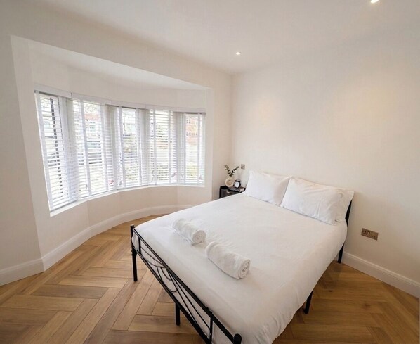 A bedroom with a bed, white linens, a nightstand with a clock, and a window with blinds.