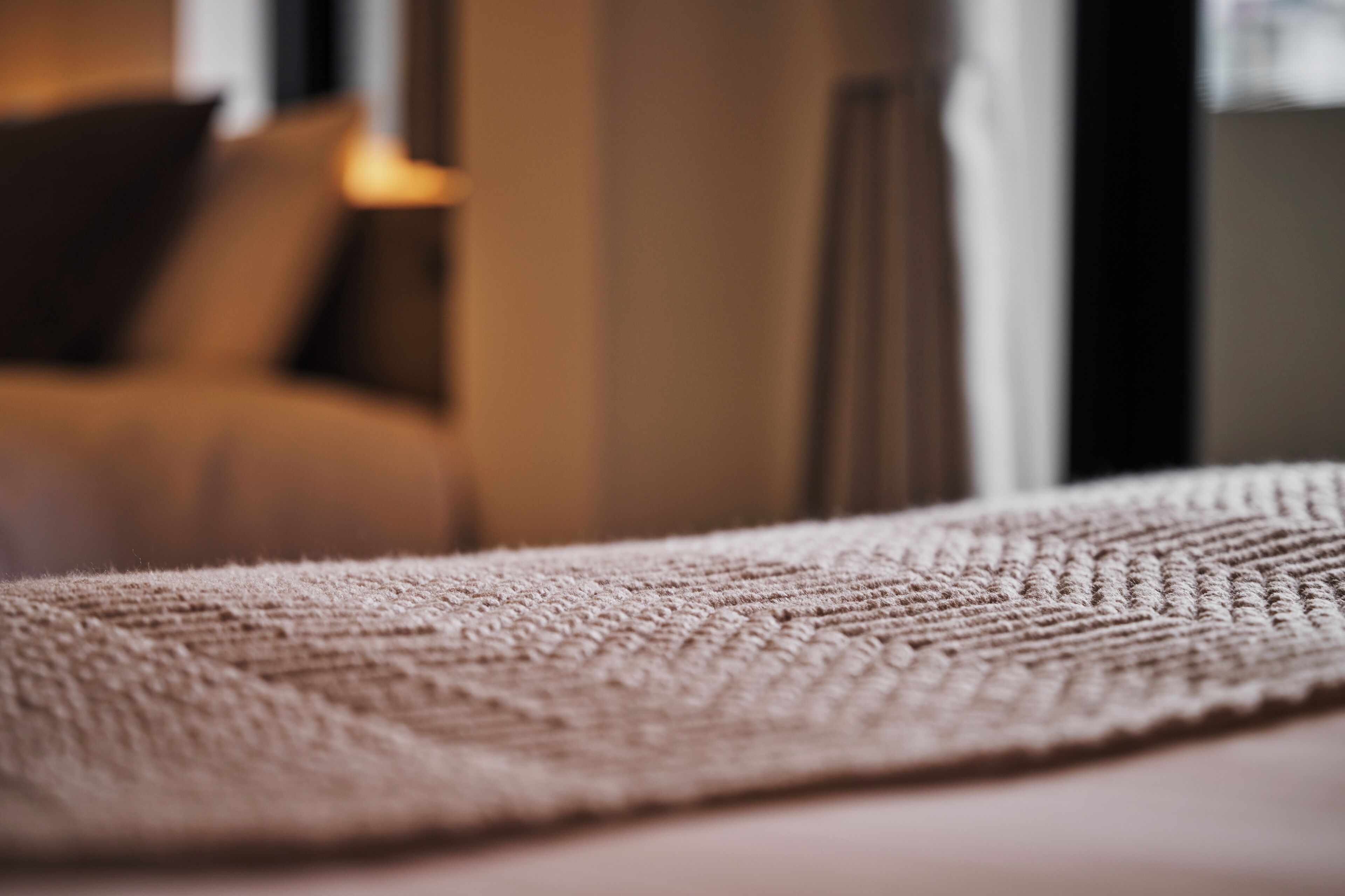 Een close-up van een textiel met een structuur, mogelijk een vloerkleed of deken, met een onscherpe achtergrond die een interieur doet vermoeden.