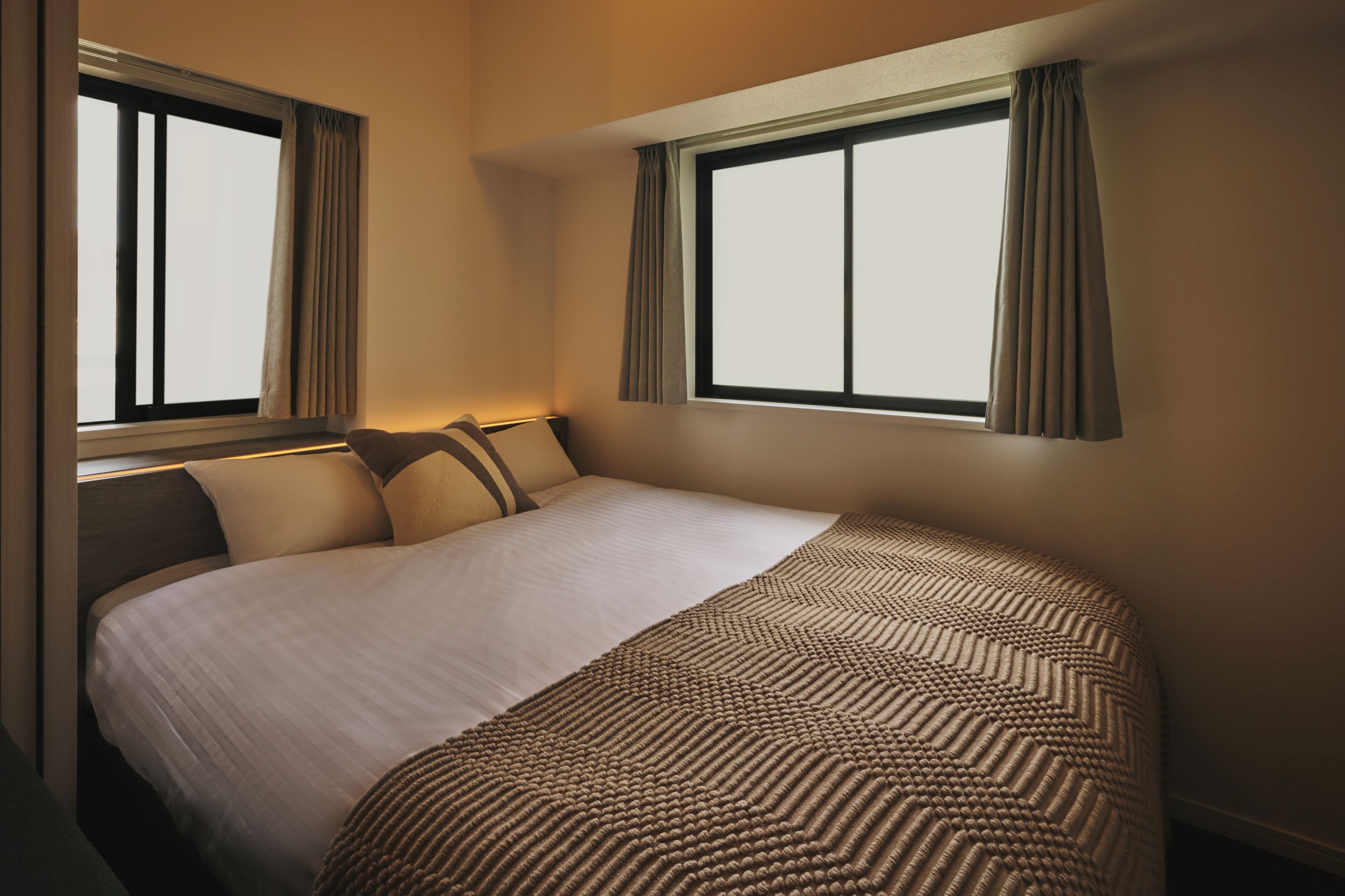 A hotel room with a large bed, two windows with curtains, and a view of the sky.