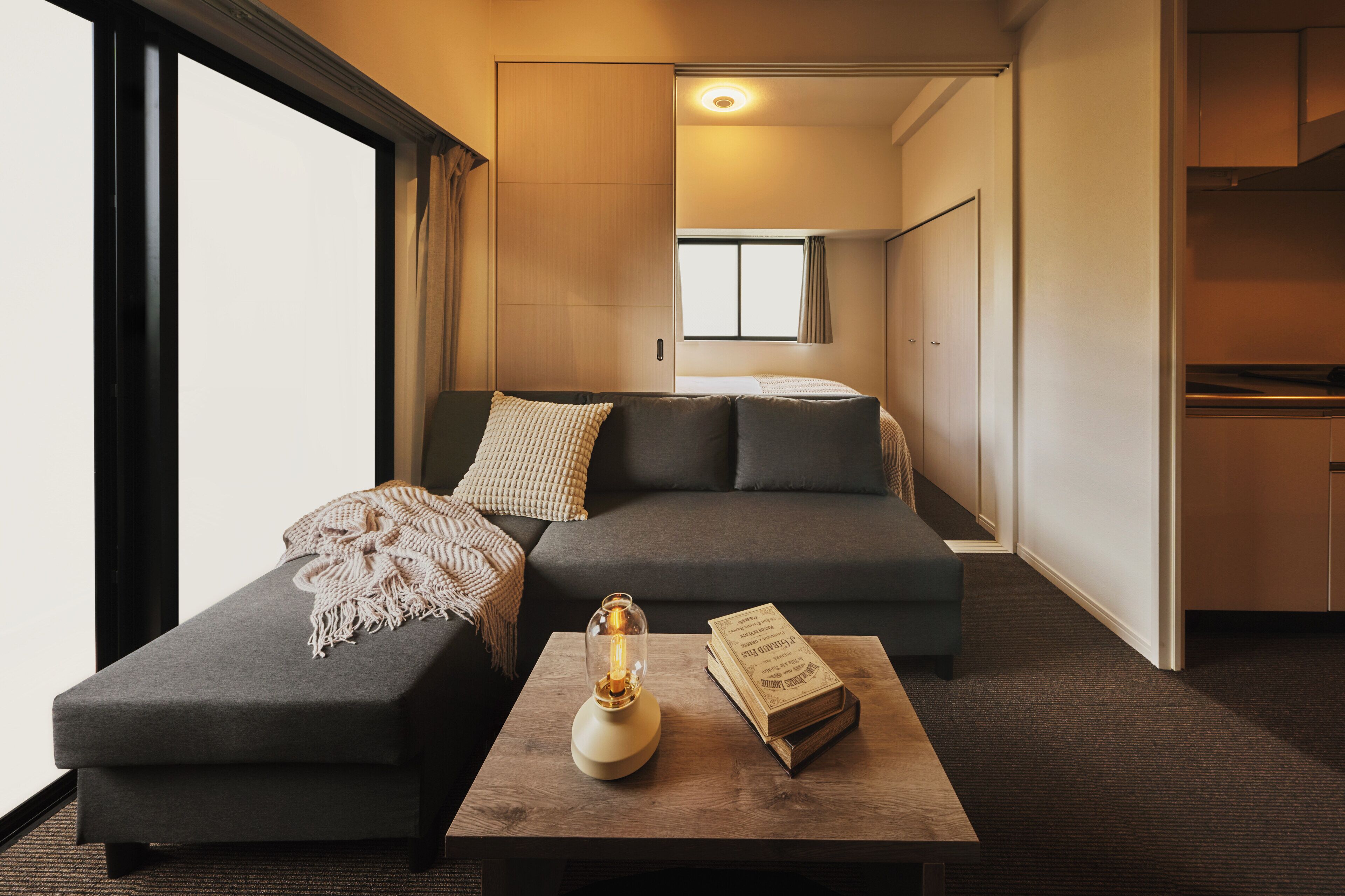 Habitación de hotel moderna con sofá, una mesa de centro con un libro y una lámpara, y vistas a la cocina y al pasillo.