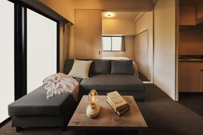 A modern hotel room with a sofa, a coffee table with a book and a lamp, and a view of the kitchen and hallway. - Residence Hotel Hakata Station2 (Fukuoka)