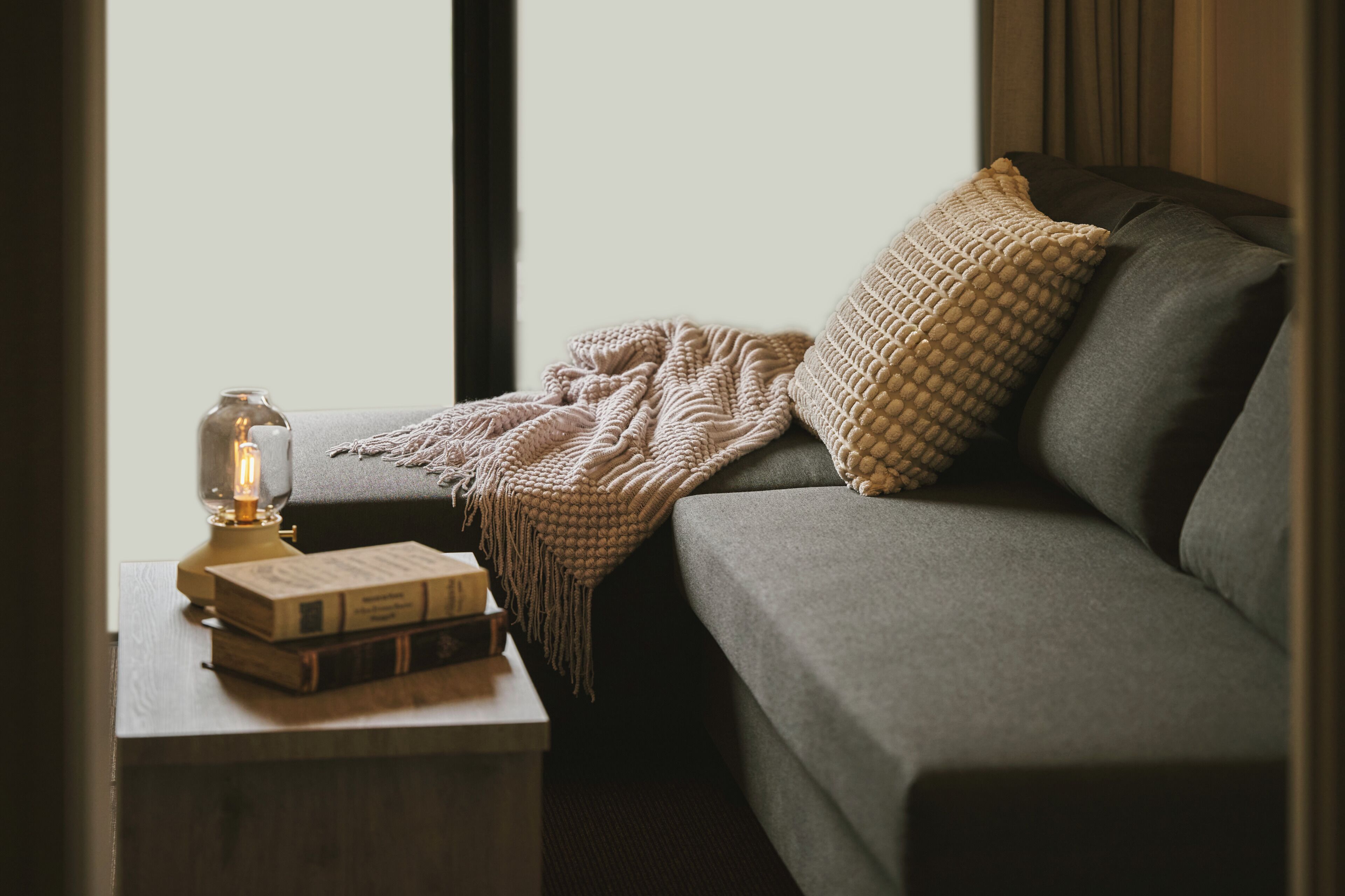 A cozy room with a grey sofa, a knitted throw blanket, a patterned pillow, a vintage-style lamp, and a stack of books on a small table.