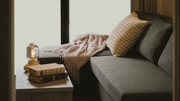 A cozy room with a grey sofa, a knitted throw blanket, a patterned pillow, a vintage-style lamp, and a stack of books on a small table.
