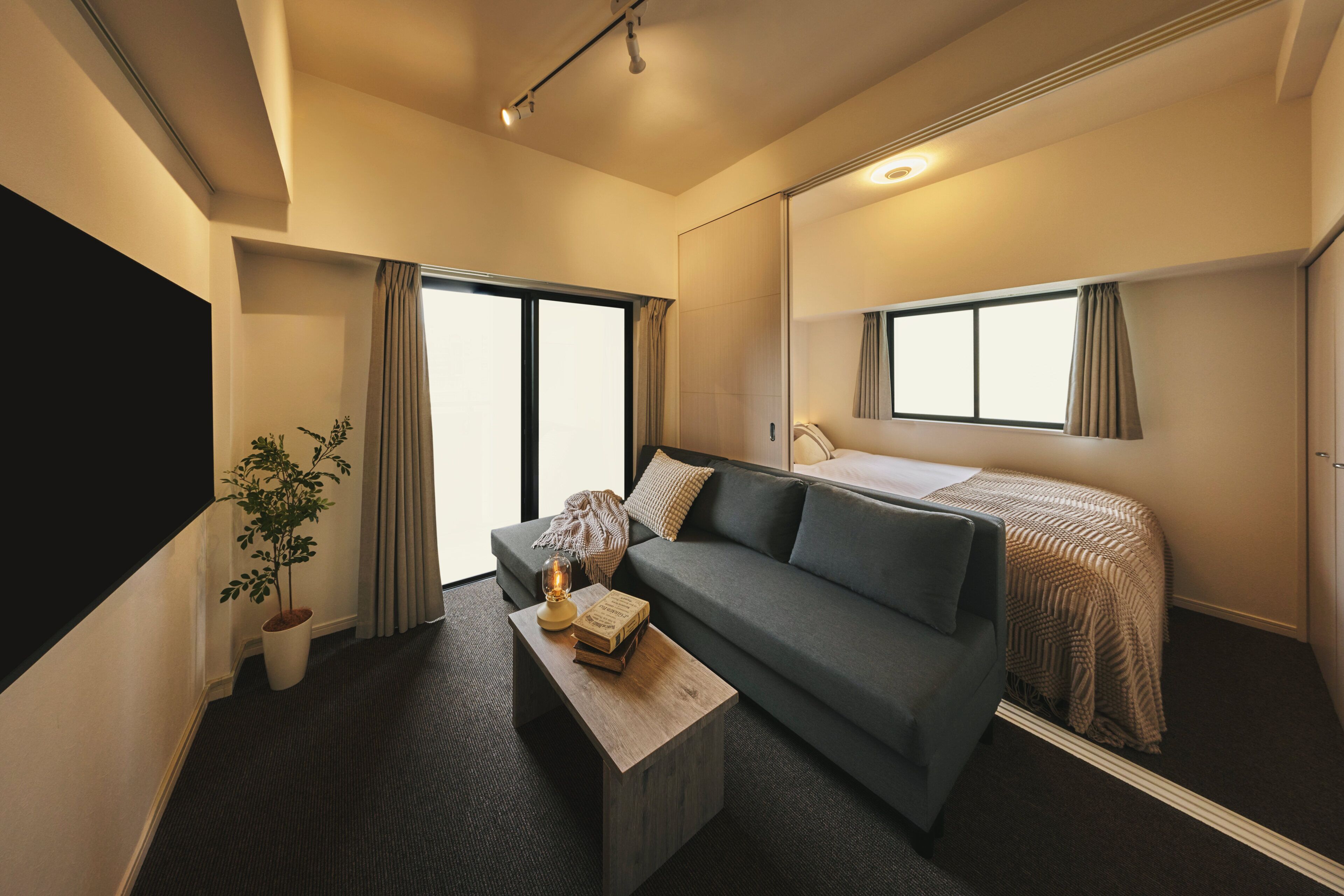 A compact hotel room with a sofa, a small table, a potted plant, and a bed with a patterned bedspread.