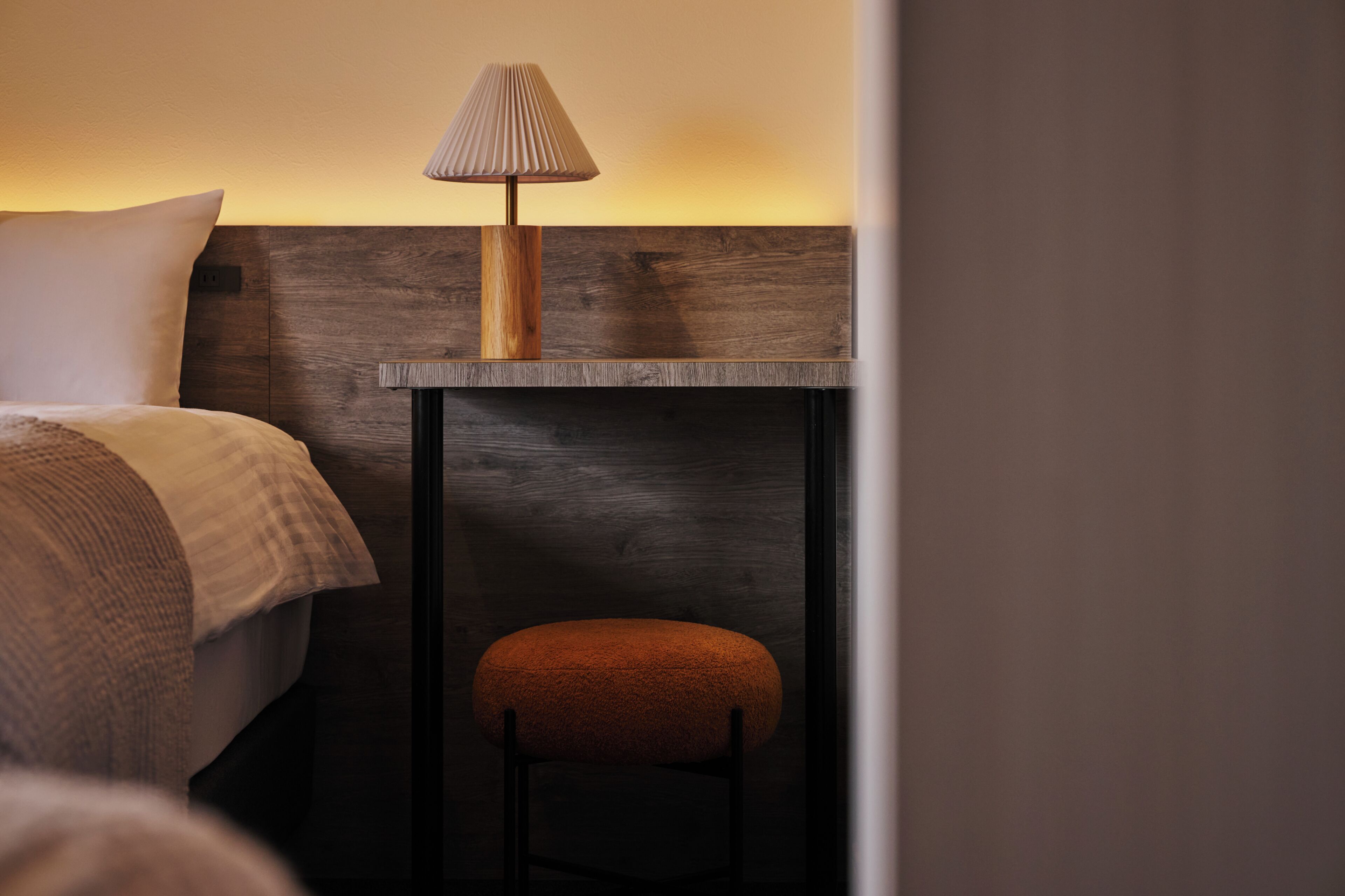 Una camera d'albergo con un letto, un comodino, una lampada e uno sgabello.