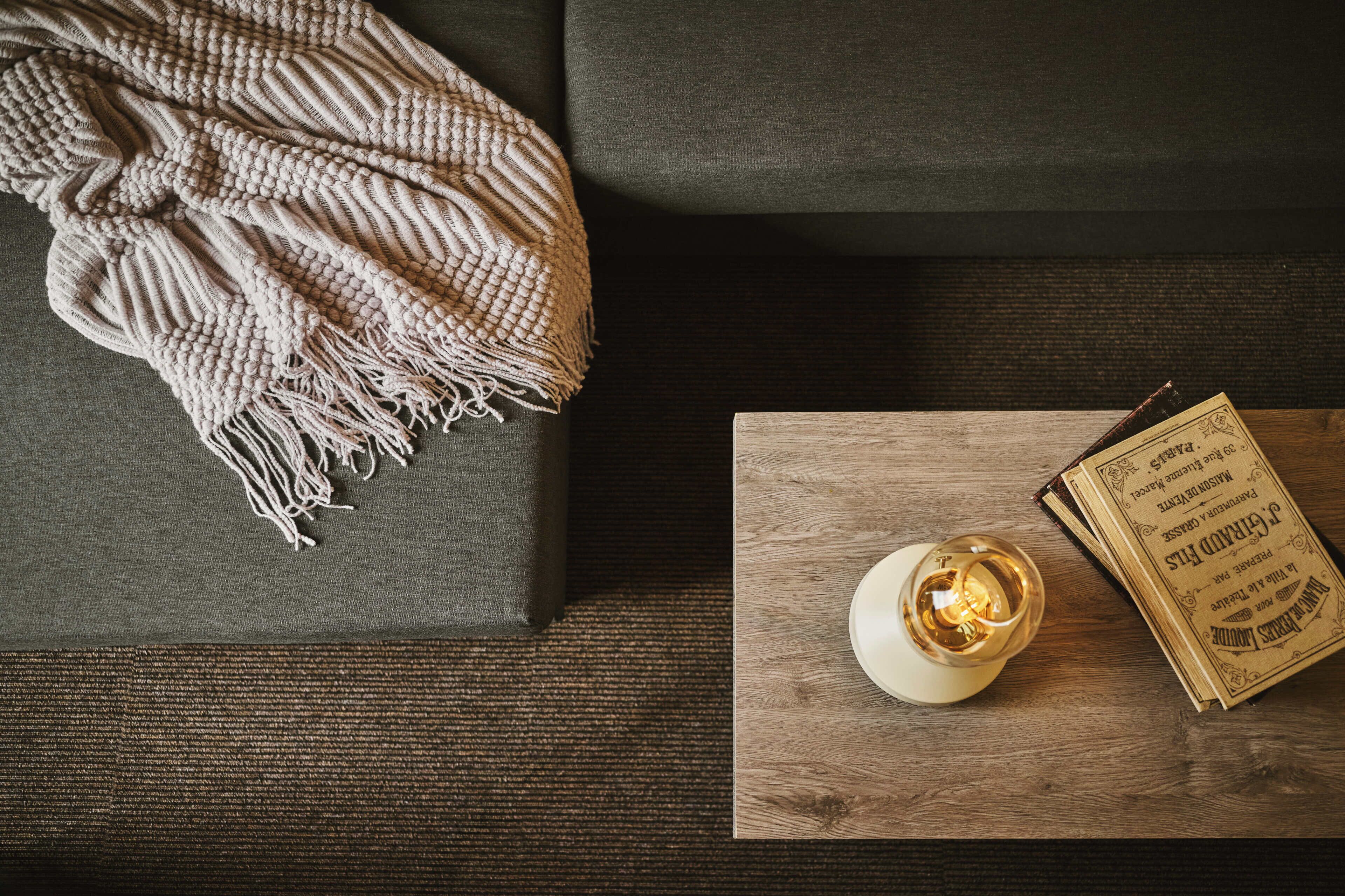 A cozy room with a grey sofa, a knitted throw blanket, a candle, and a vintage book on a wooden side table.