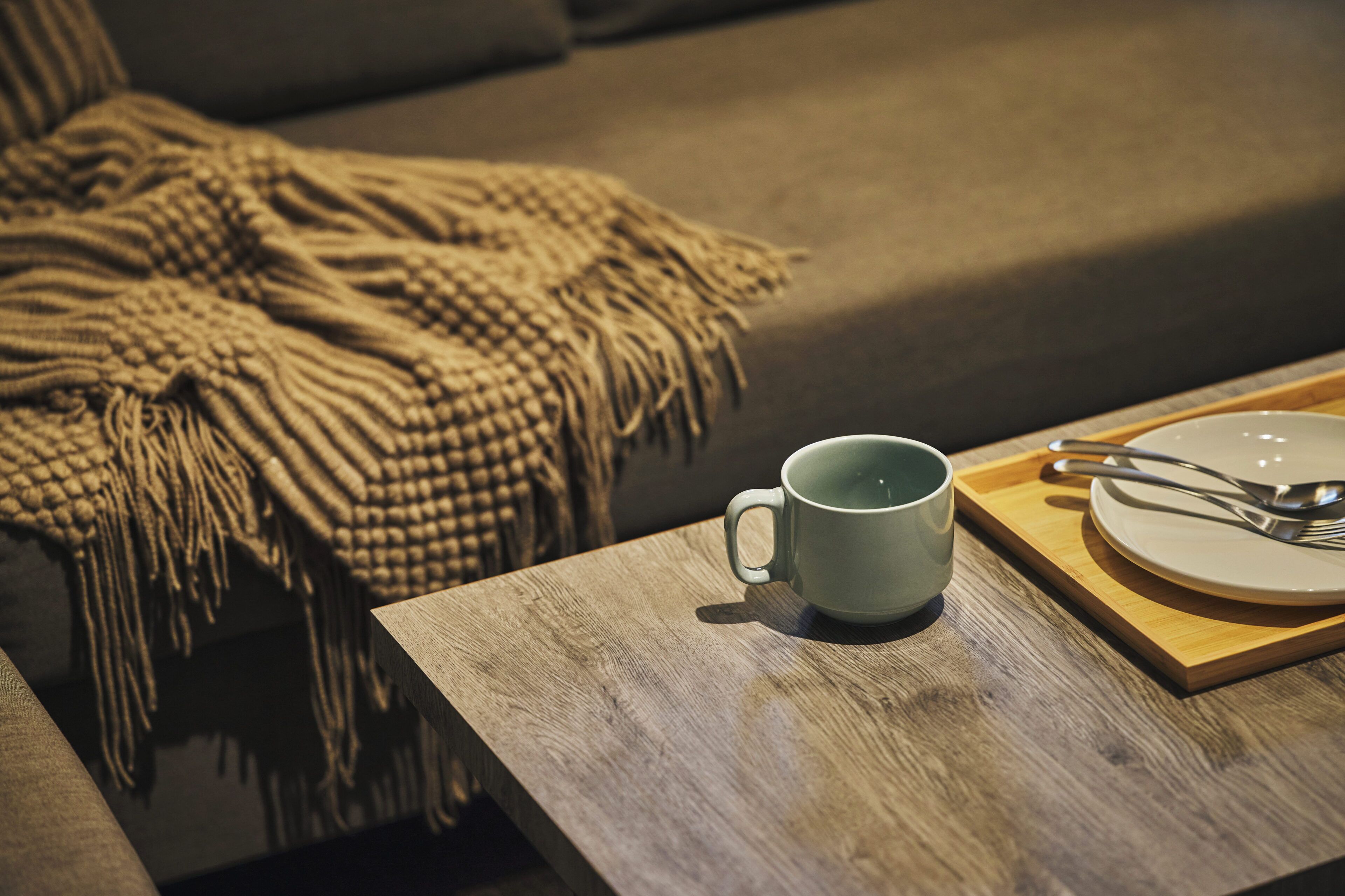 A wooden table with a mug and a plate with cutlery, a beige blanket on a couch, and a wooden tray.