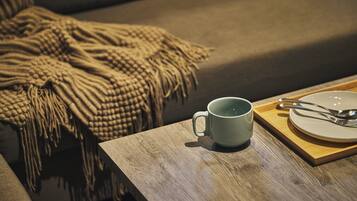 A wooden table with a mug and a plate with cutlery, a beige blanket on a couch, and a wooden tray.