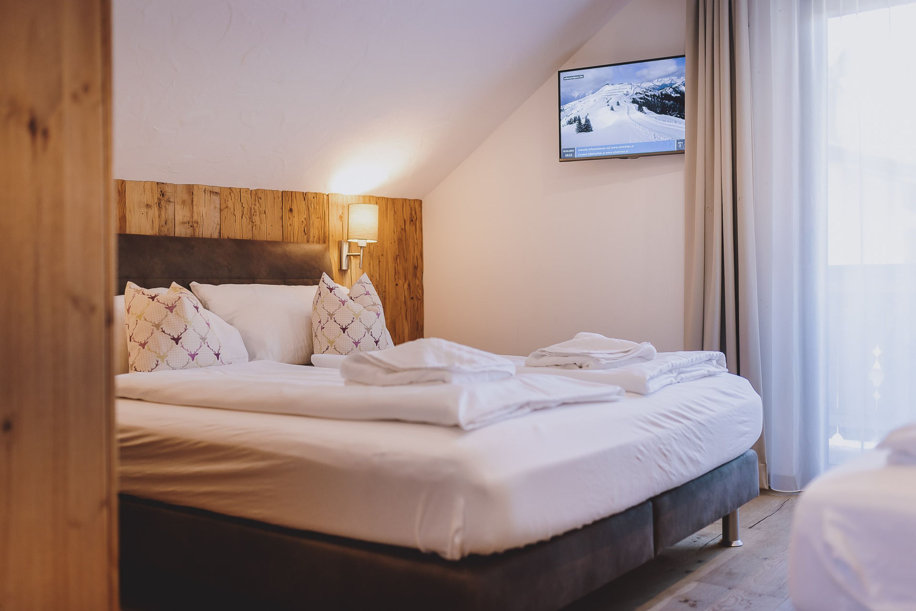Ein ordentlich bezogenes Bett mit weißen Leinen, einem hölzernen Kopfteil und einem an der Wand befestigten Fernseher, der eine verschneite Landschaft zeigt.