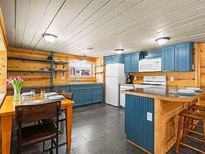 A kitchen with wooden cabinets, a central island, and a dining area with chairs and a table set for two. - Quaint 3-bedroom Boyne City Cabin/ 3-Minutes to Boyne Mountain (Boyne City)