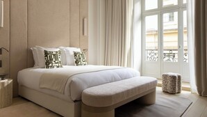 Quarto de hotel com uma cama grande, um banco, um banquinho pequeno, uma mesinha de cabeceira e uma janela com cortinas.