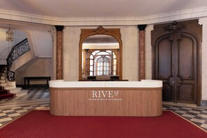 Ein Empfangstresen mit der Beschriftung 'RIVE 15 APARTMENTS PARISIENS', eine imposante Treppe und eine große Holztür.