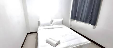 A single bed with white linens, a dark headboard, and a window with grey curtains.