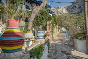 A narrow stone pathway flanked by colorful pots and potted plants, leading to a building with a stone facade and a mountain backdrop. - Villa 'Lila' with Sea View, Wi-Fi and Air Conditioning (Órmos Aiyialís)