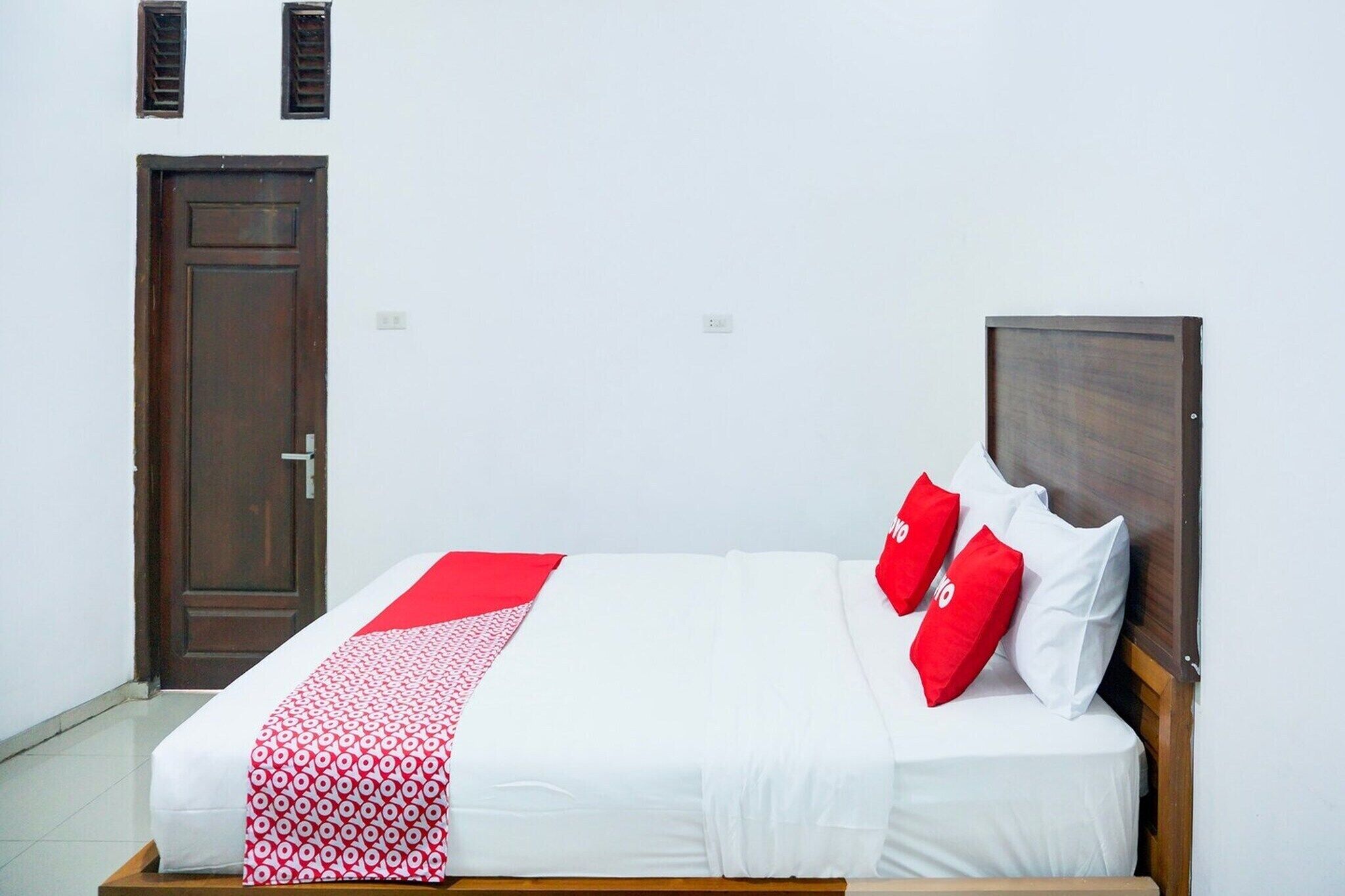 A bed with a white and red bedspread, two red pillows, a wooden headboard, and a dark wooden door.
