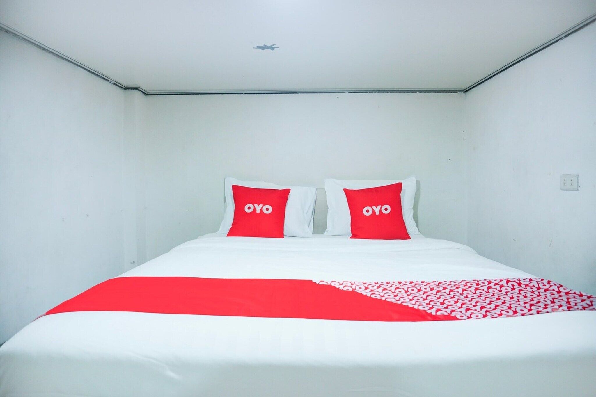 Un lit simple avec une literie blanche et rouge, deux oreillers portant le logo « OYO », et une couverture à motifs rouges et blancs.
