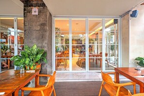 Een eetruimte met houten tafels en stoelen, een grote potplant en een uitzicht door glazen deuren op een zitgedeelte buiten.