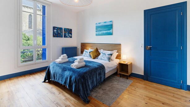 Une chambre avec une grande fenêtre, un lit avec une literie bleue, une tête de lit en bois, une table de chevet et une porte bleue.