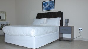 Ein ordentlich bezogenes Bett mit weißen Leinen, ein Nachttisch mit Schublade, eine Wandleuchte und ein gerahmtes Bild über dem Bett.