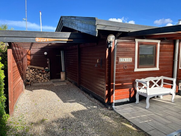 A wooden cabin with a sign reading 'Berglyck 6' and 'UTKIEK' on the door, a bench outside, and a gravel path leading to the entrance.