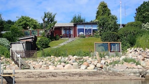 A lakeside property with a wooden dock, a red building with large windows, and a stone pathway leading to the house. - fewo1846 - Haus Utkiek - fewo1846 - Utkiek / Three-bedroom vacation home (Glücksburg (Ostsee))