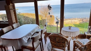 A dining area with a table and chairs, a round table with a decorative item, and a view of the sea through large windows. - fewo1846 - Haus Utkiek - fewo1846 - Utkiek / Three-bedroom vacation home (Glücksburg (Ostsee))