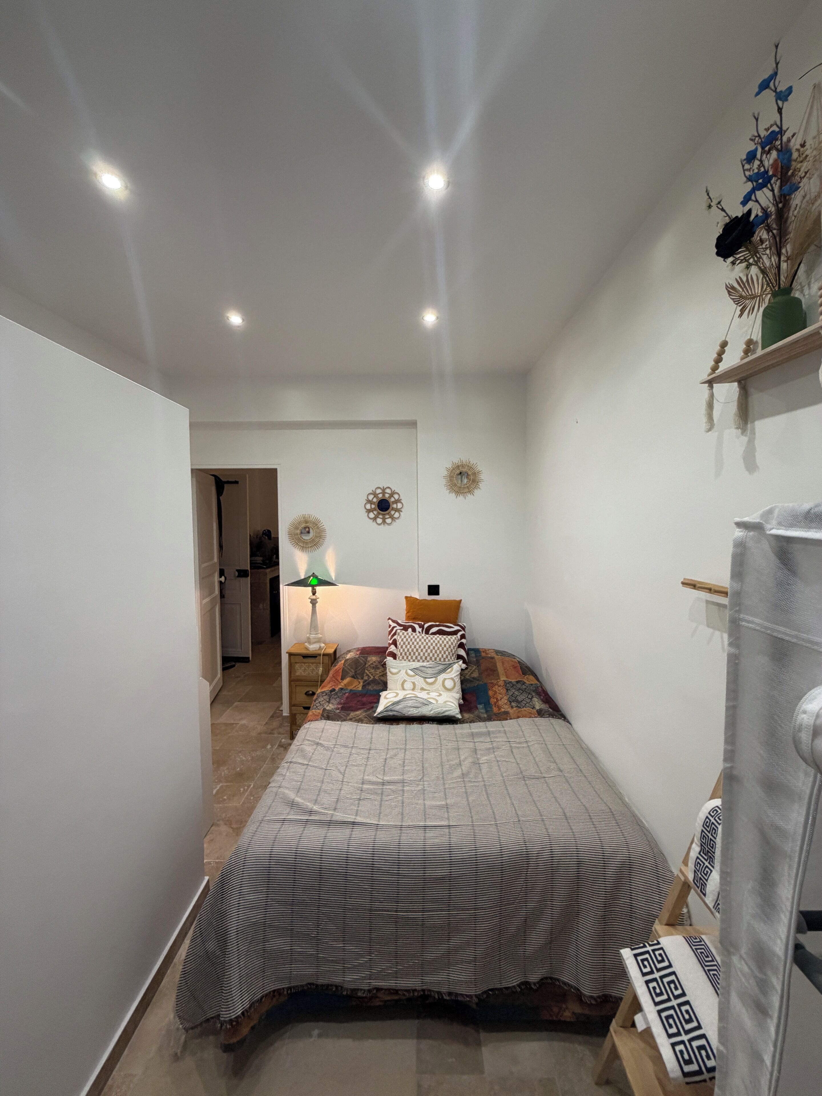 A narrow hallway leading to a bedroom with a bed, a nightstand, and a wall-mounted shelf with decorative items.