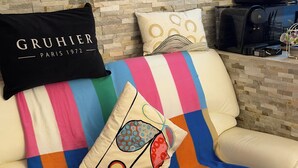Un canapé en cuir beige agrémenté de couvertures rayées colorées et de coussins décoratifs, une table basse en verre sur laquelle repose un objet décoratif, et un mur dont le motif rappelle la pierre.