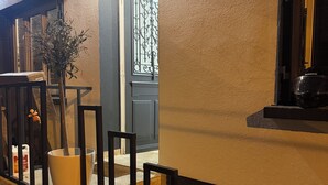 Un escalier menant à une porte surmontée d’un portail en fer forgé décoratif, avec un pot de fleurs et un sac posé sur les marches.