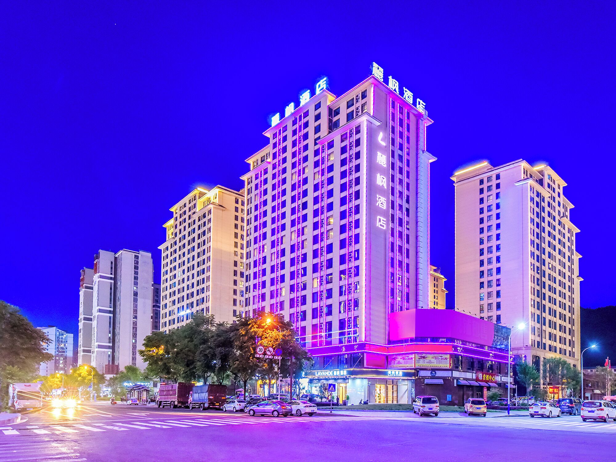 Un hotel de varios pisos con señalización iluminada, una entrada destacada y vistas a la calle con vehículos y árboles.