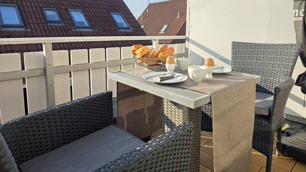 Un balcone con mobili in vimini, un tavolo con prodotti per la colazione e tetti in tegole rosse sullo sfondo.