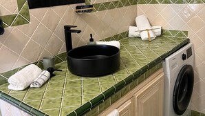 Un baño con encimera de azulejos verdes, un lavabo negro, un espejo, un perchero y una lavadora.