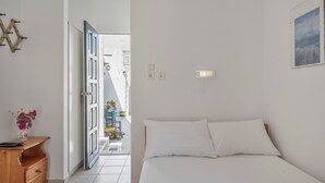 Une chambre à coucher comprenant un lit, une commode en bois, une lampe fixée au mur et une porte donnant sur un balcon.