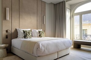 Habitació d'hotel moderna amb un llit gran, una taula de nit, una banca al costat de la finestra i vista a la ciutat.