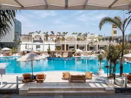 A pool area with lounge chairs and umbrellas, a clear sky, and a building with a sign reading 'Taga Arabia'.