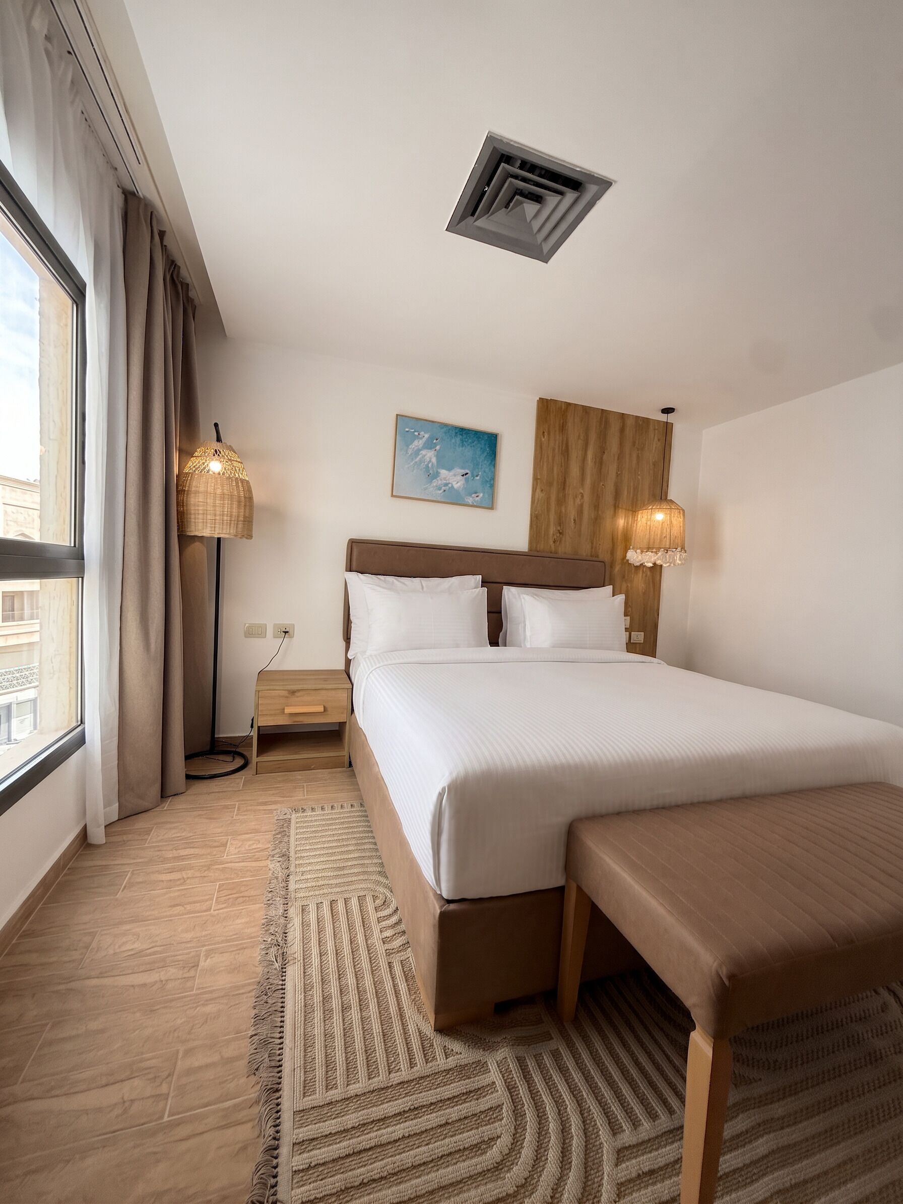 A hotel room with a large bed, a bedside table, a lamp, a painting, and a view of the outside.