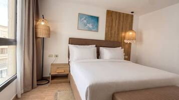 A hotel room with a large bed, a bedside table, a lamp, a painting, and a view of the outside.