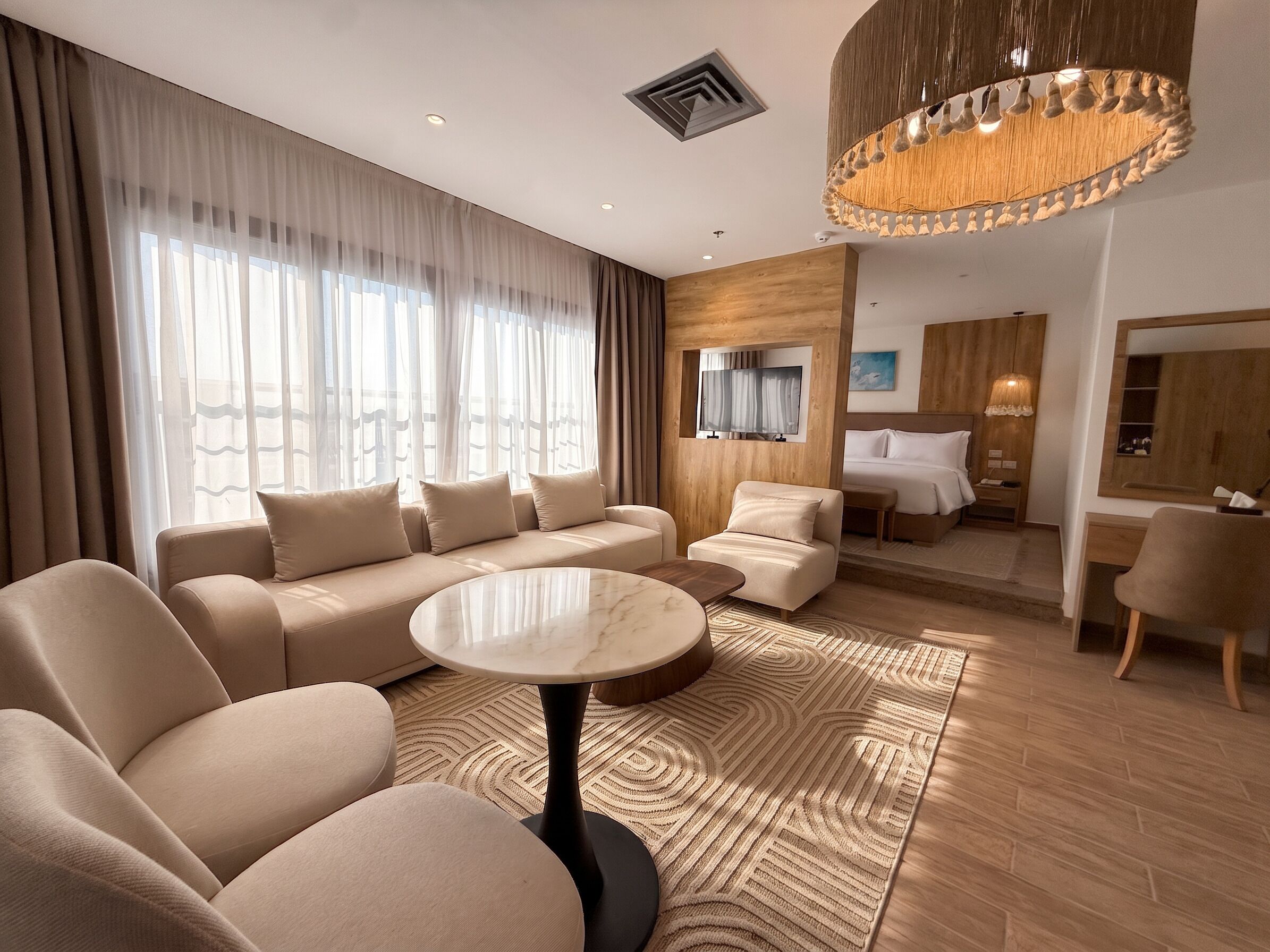 Una habitación de hotel moderna con sofá, una mesa redonda, cama, escritorio y un ventanal con cortinas.