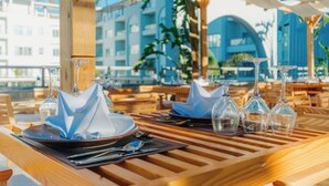Une table dressée pour le dîner, avec de la vaisselle et des verres transparents, des meubles en bois et, en arrière-plan, une vue sur un bâtiment bleu.