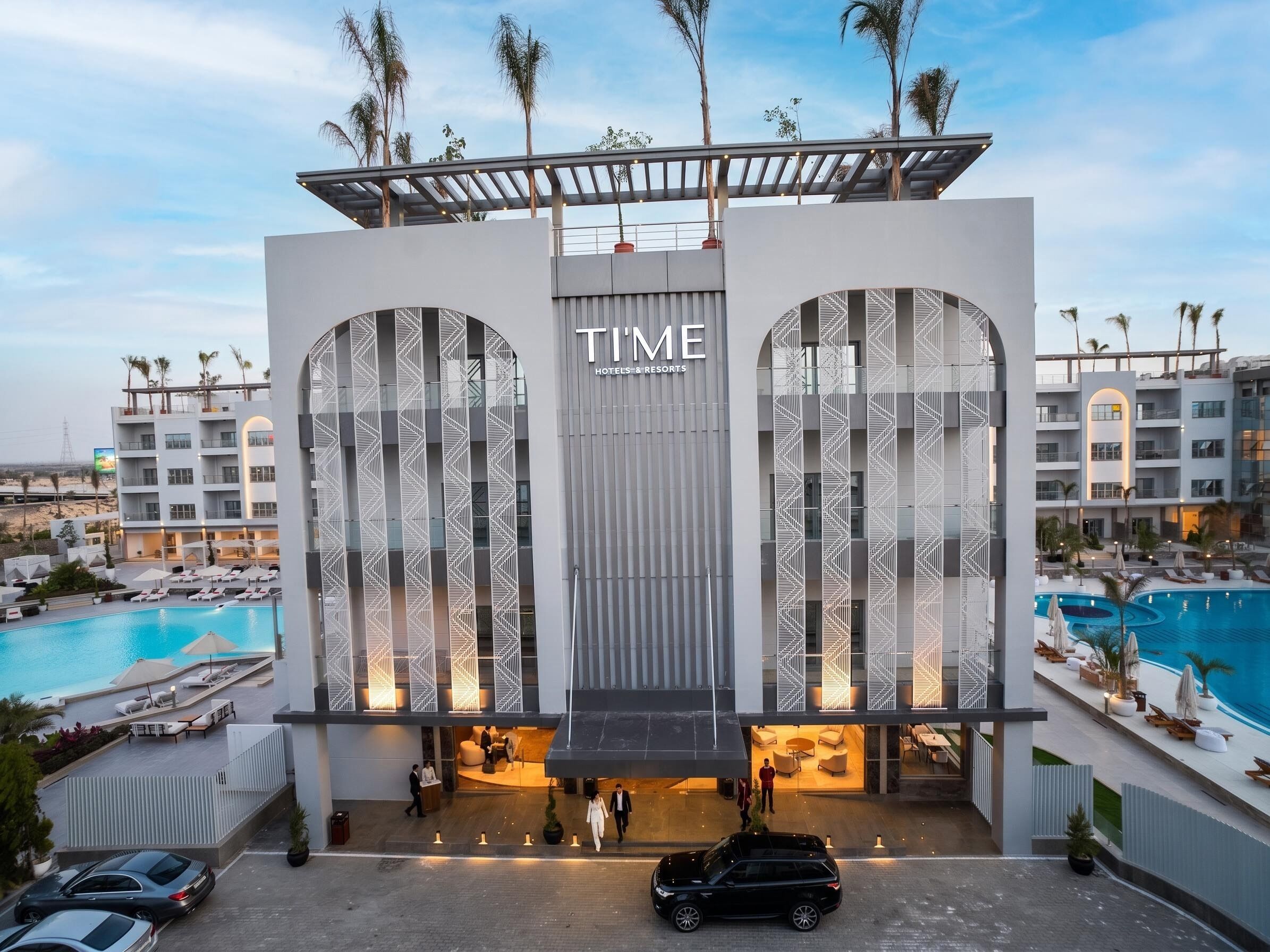 A hotel with a modern design, featuring a large sign that reads 'TIME HOTELS & RESORTS'. There are multiple floors visible, and the hotel has a pool area with lounge chairs.