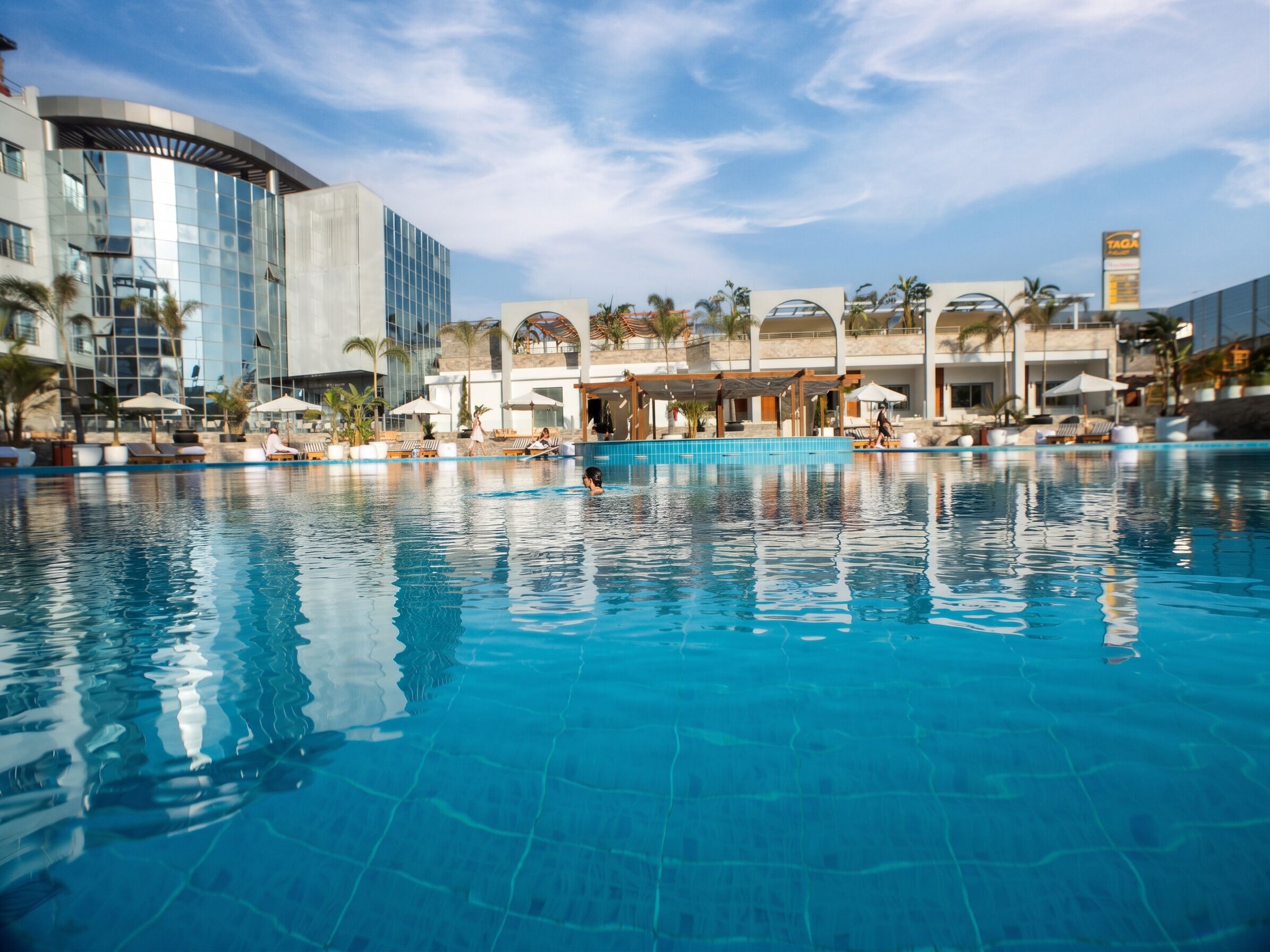 A swimming pool with lounge chairs and umbrellas, surrounded by modern buildings and palm trees.