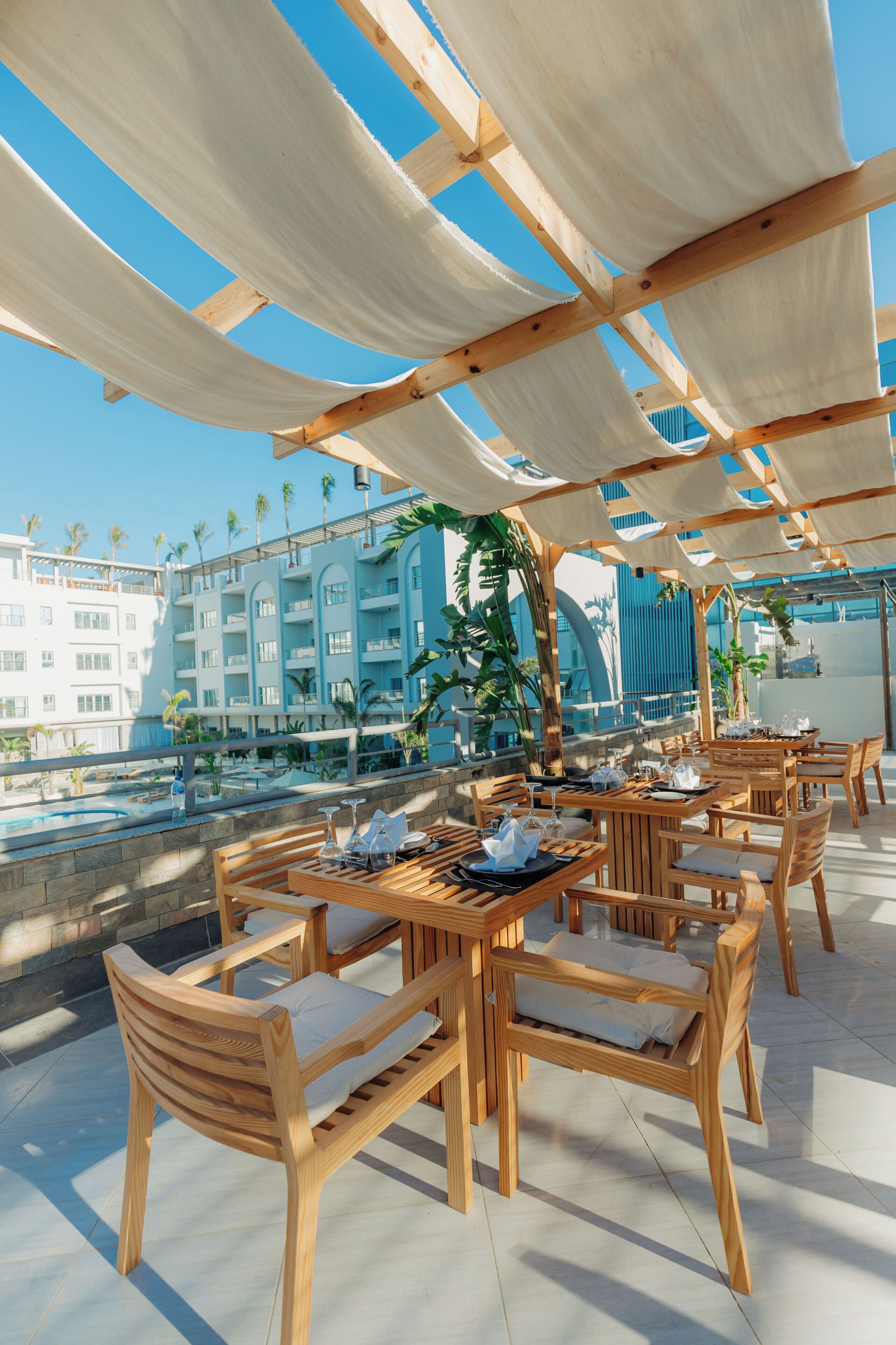 An outdoor dining area with wooden tables and chairs, a pool in the background, and a view of a hotel building.