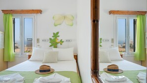 A hotel room with a bed, pillows, a hat, a green bedspread, a wall decoration, and a view of the sea. - Room 6 at Pagali Hotel in Amorgos (Órmos Aiyialís)